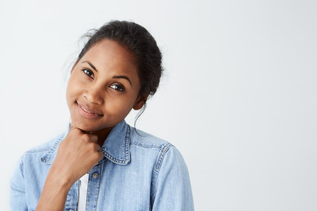 Nahaufnahme der afroamerikanischen Frau mit den dunklen warmen Augen und dem ansprechenden Lächeln gekleidet in der Freizeitkleidung, die auf weißer Wand aufwirft. Hübsche dunkelhäutige Frau, die glücklichen Blick über Weiß isoliert hat.