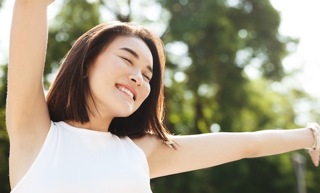 Nahaufnahme der asiatischen Frau, die Hände nach oben streckt und lächelt, im Park spazieren geht, sorglos und glücklich aussehend