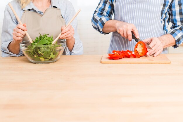 Nahaufnahme der Frau den Salat und ihren Ehemann vorbereitend, die den grünen Pfeffer auf Holztisch schneiden