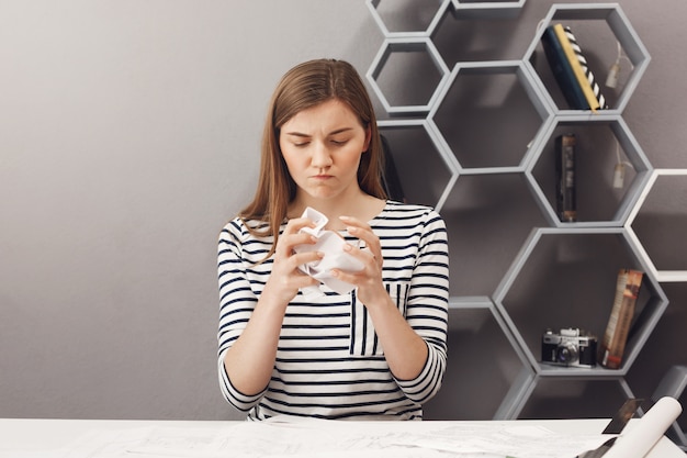 Nahaufnahme der frustrierten schönen europäischen dunkelhaarigen freiberuflichen Designerin zerknittert Papier mit Händen und wütendem Gesichtsausdruck, wütend zu sein, dass nichts klappt.