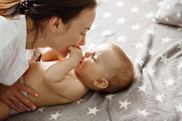 Kostenloses Foto nahaufnahme der glücklichen gut aussehenden mutter, die mit ihrem neugeborenen sohn spielt, seine kleinen hände küsst und sanft beißt. glückliches mutterschaftskonzept.