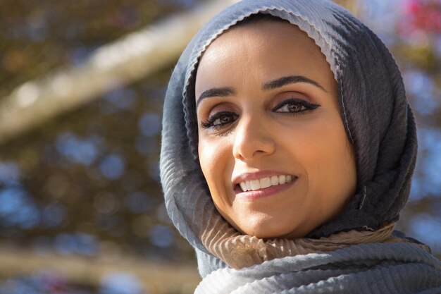 Nahaufnahme der glücklichen muslimischen Frau. Hübsche junge Frau im grauen Hijab mit gutem Make-up, das die Kamera lächelt, die im Park auf Baumhintergrund steht. Schönheits- und Glückskonzept