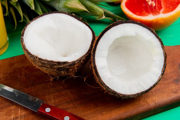 Nahaufnahme der halb geschnittenen Kokosnuss und des Messers auf Schneidebrett mit anderen Zitrusfrüchten auf grünem Hintergrund
