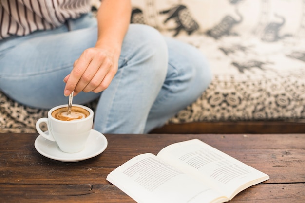 Nahaufnahme der Hand der Frau, die Kaffee Latte mit Löffel und Buch auf Tabelle rührt