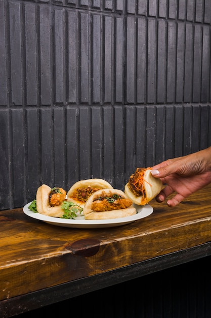 Nahaufnahme der Hand der Frau Gua Bao auf weißer Platte über dem Holztisch nehmend