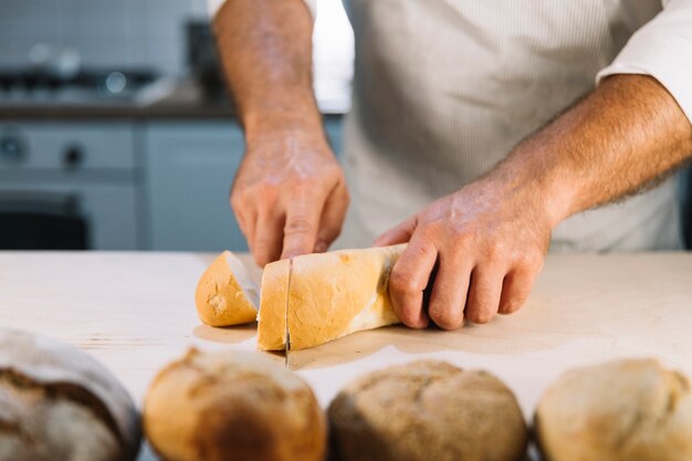 Nahaufnahme der Hand des männlichen Bäckers, die Brot mit Messer auf Küchenarbeitsplatte schneidet
