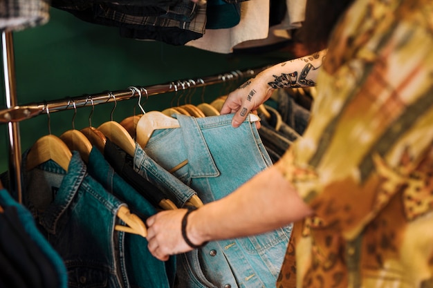 Nahaufnahme der hand des mannes die blaue jacke wählend, die an der schiene im kleidungsshop hängt