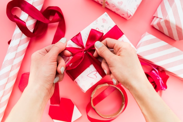 Nahaufnahme der Hand einer Frau, die das rote Band auf Geschenkbox über dem rosa Hintergrund bindet