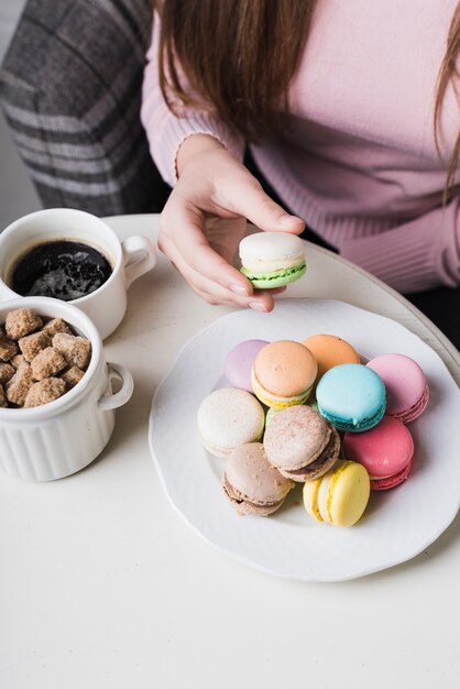 Nahaufnahme der Hand einer Frau, die Makrone mit Würfel des Kaffees und des braunen Zuckers in der Schale auf Tabelle hält