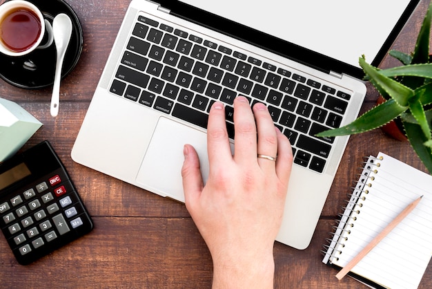 Nahaufnahme der Hand einer Person, die auf Laptop mit Taschenrechner schreibt; Kaffeetasse und Spiralblock mit Bleistift auf Schreibtisch aus Holz