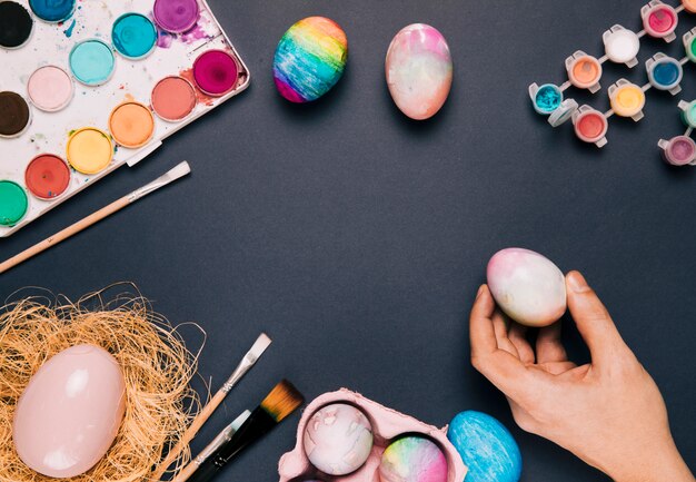 Nahaufnahme der Hand einer Person, die das gemalte Ei mit Farbenfarbe und -pinseln auf schwarzem Hintergrund hält