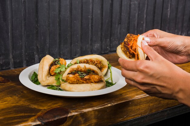 Nahaufnahme der Hand einer Person, die Gua Bao von der weißen Platte auf Holztisch nimmt