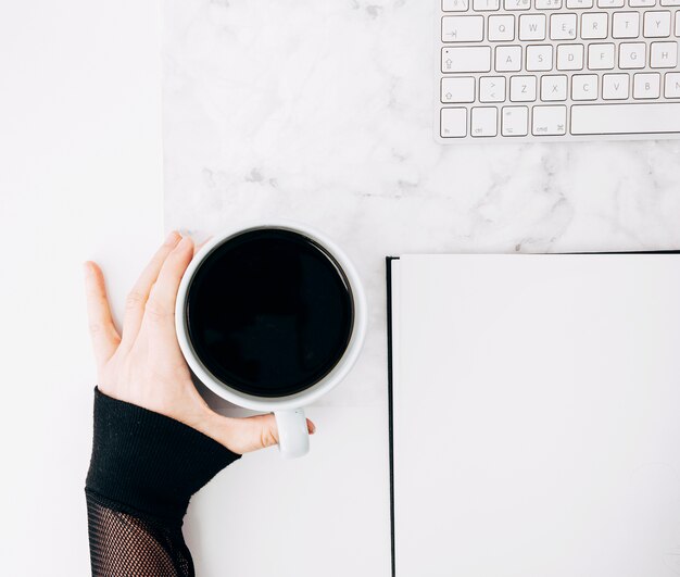 Nahaufnahme der Hand einer Person, die schwarze Kaffeetasse mit Tagebuch und Tastatur auf Schreibtisch hält