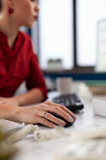 Nahaufnahme der Hand mit drahtloser Computermaus im Startup-Büro. Unternehmerfinger mit Ringkliken am Schreibtisch. Manager im roten Hemd, der Inhalte auf dem Desktop-Computer im Arbeitsbereich des Unternehmens scrollt.