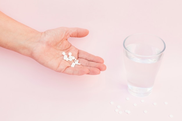 Nahaufnahme der Hand mit weißen Tabletten und Glas Wasser auf rosa Hintergrund