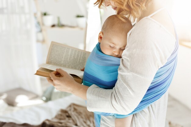 Nahaufnahme der jungen Mutter, die Märchen für ihren neugeborenen kleinen Sohn im bequemen hellen Schlafzimmer liest. Baby schläft ein, während sie las.