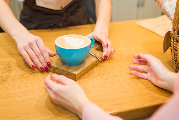 Nahaufnahme der Kellnerin dem Kunden auf Holztisch Kaffeetasse mit Lattekunstschaum gebend