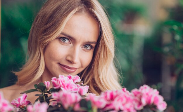 Nahaufnahme der lächelnden blonden jungen Frau mit den rosa Blumen, die Kamera betrachten