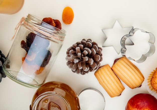 Nahaufnahme der Rosinen, die aus Glas und Tannenzapfen mit Pfirsichmarmelade und Keksen auf weißem Tisch verschüttet werden