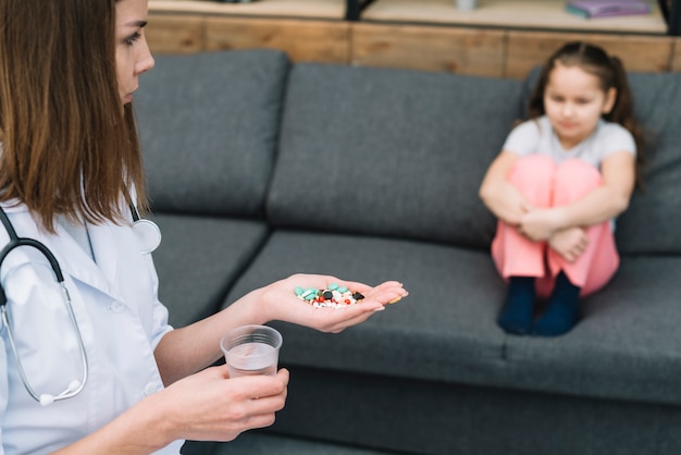 Nahaufnahme der Ärztin Medizin und Glas Wasser halten das Mädchen betrachtend, das auf Sofa sitzt