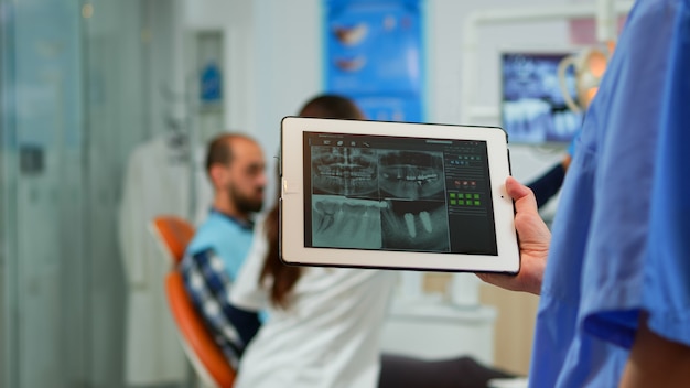 Kostenloses Foto nahaufnahme der zahnarztkrankenschwester, die tablet mit digitaler radiographie hält, während der arzt mit dem patienten im hintergrund arbeitet und das zahnproblem untersucht, das auf einem stomatologischen stuhl in der zahnklinik sitzt.