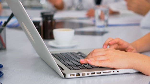 Kostenloses Foto nahaufnahme des assistenten mit laptop, der sitzungsinformationen schreibt, während ärzte im hintergrund während der medizinischen konferenz am schreibtisch im krankenhausbüro diskutieren. professionelles brainstorming im team