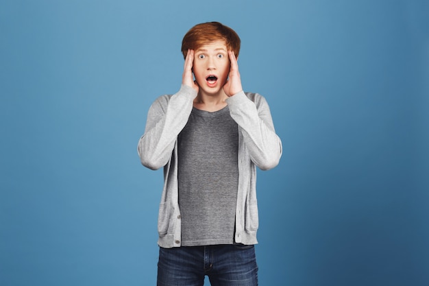 Nahaufnahme des ausdrucksstarken jungen gutaussehenden Teenagers mit roten Haaren im lässigen Outfit, das den Kopf mit den Händen drückt und schockiert ist, den besten Freund mit seiner Freundin zu sehen.