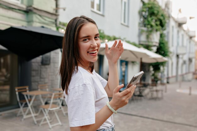 Nahaufnahme des Außenfotos einer charmanten, süßen Frau mit wunderbarem Lächeln, die auf dem Smartphone scrollt und ihr Haar auf der Straße der Stadt berührt