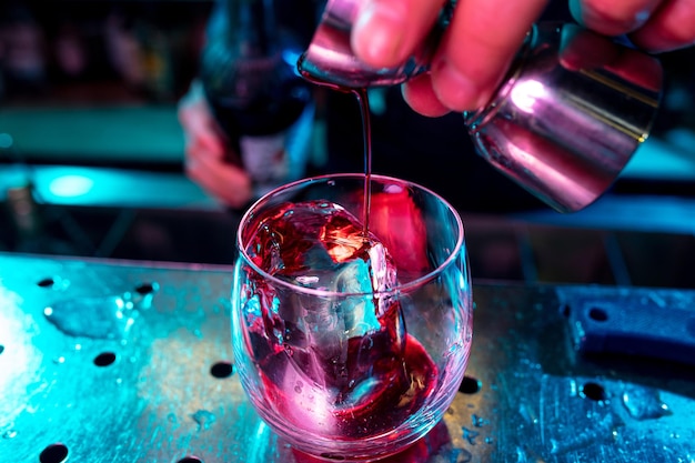 Nahaufnahme des Barmanns, der einen alkoholischen Cocktail mit Schuss in mehrfarbigem Neonlicht zubereitet