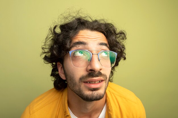 Nahaufnahme des beeindruckten jungen gutaussehenden kaukasischen Mannes, der das Tragen der Brille lokalisiert auf olivgrüner Wand nach oben schaut