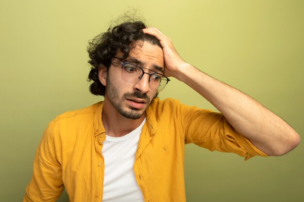 Nahaufnahme des besorgten jungen gutaussehenden Mannes, der die Brille trägt, die Hand auf Kopf setzt, der lokalisiert auf olivgrüne Wand schaut