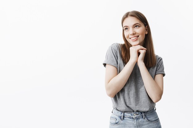 Nahaufnahme des charmanten gutaussehenden jungen Studentenmädchens mit den dunklen Haaren und den Augen im lässigen Outfit lächelnd, mit verträumtem und glücklichem Gesichtsausdruck beiseite schauend, Hände zusammen unter Gesicht haltend