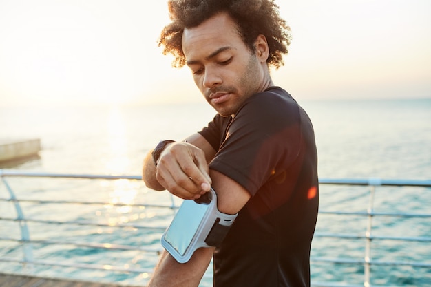 Nahaufnahme des dunkelhäutigen männlichen Athleten, der mobile Armtasche repariert. Morgen Outdoor-Training hinter dem Meer. Sport-, Technologie- und Freizeitkonzept.