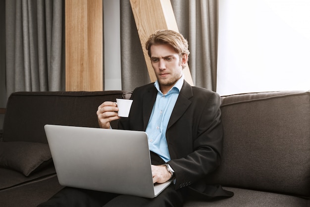 Nahaufnahme des eleganten unrasierten Mannes, der Kaffee trinkt, im Laptop-Monitor mit ernstem und unbefriedigtem Ausdruck schaut und von zu Hause aus arbeitet.
