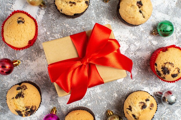 Nahaufnahme des Geschenks mit rotem Band zwischen frisch gebackenen, köstlichen kleinen Cupcakes und Dekorationszubehör auf der Eisoberfläche