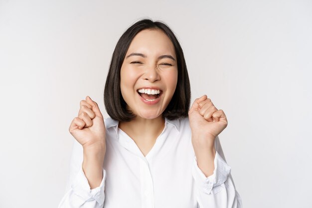 Nahaufnahme des Gesichtsporträts einer tanzenden asiatischen Frau, die triumphierend lächelt und mit fröhlichen Emotionen feiert, die über weißem Hintergrund stehen. Kopieren Sie Platz