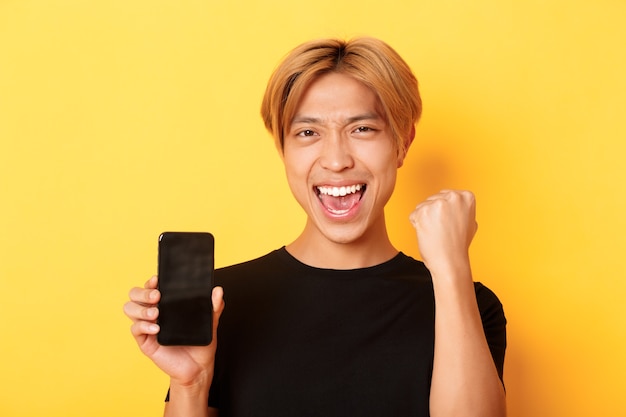 Nahaufnahme des glücklichen fröhlichen asiatischen Kerls, der Smartphonebildschirm zeigt und ja sagt, Faustpumpe als triumphierend, gewinnend oder Ziel, gelbe Wand