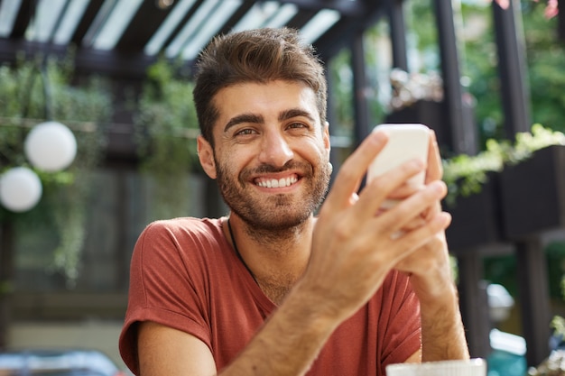 Nahaufnahme des glücklichen gutaussehenden jungen Mannes lächelnd, während draußen Café sitzen und Handy verwenden