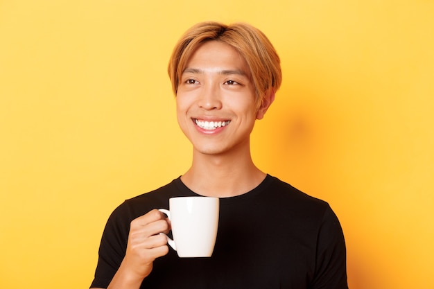 Nahaufnahme des glücklichen hübschen jungen asiatischen Kerls mit blondem Haar, träumerisch aussehend und lächelnd, während Kaffee oder Tee trinkend, über gelber Wand stehend.