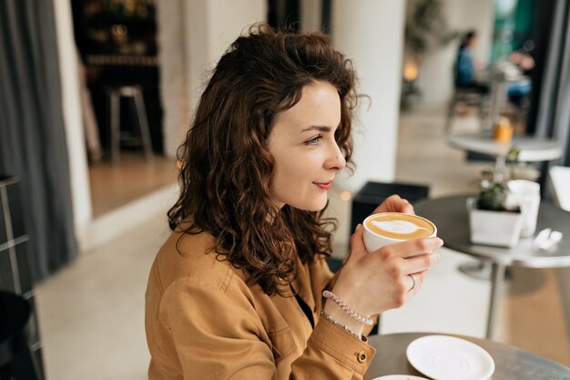 Nahaufnahme des Innenporträts einer hübschen, charmanten Dame mit lockigem Haar, die ein braunes Hemd trägt, hält eine Tasse mit Kaffee und freut sich auf die Erholung am Morgen