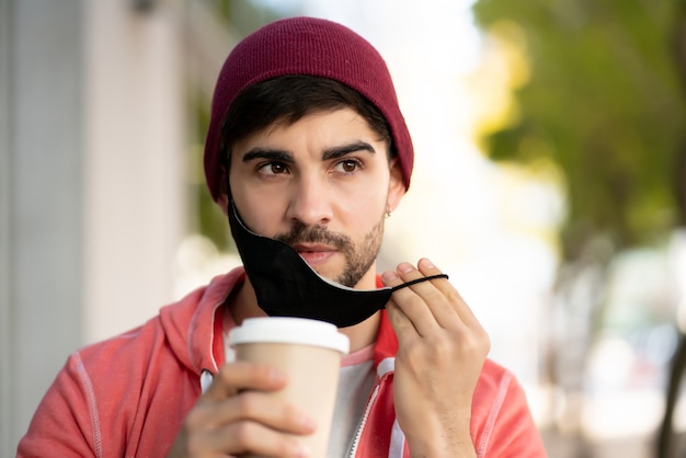 Nahaufnahme des jungen Mannes, der eine Schutzmaske trägt und Kaffee trinkt, während draußen an der Straße steht