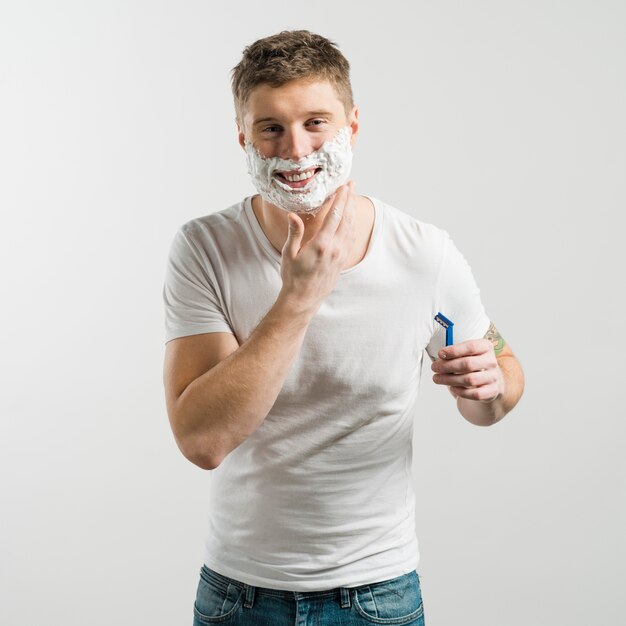 Nahaufnahme des lächelnden jungen Mannes mit dem Rasieren des Schaums auf seinem Gesicht, das in der Hand Rasiermesser zur Kamera schaut