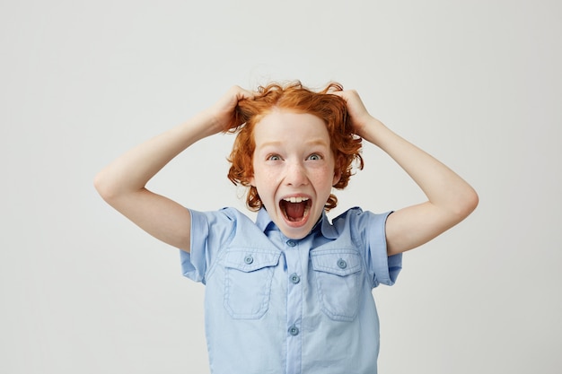 Nahaufnahme des lustigen kleinen Jungen mit roten Haaren und Sommersprossen, die Haare mit den Händen ziehen und mit überraschtem Ausdruck schreien