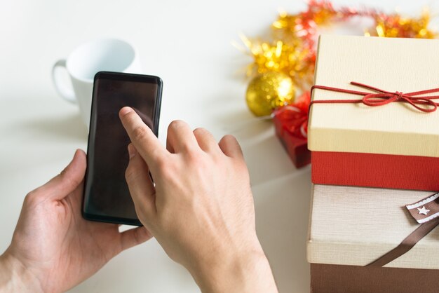 Nahaufnahme des Mannes, der bei Tisch Smartphone mit Weihnachtsgeschenken verwendet