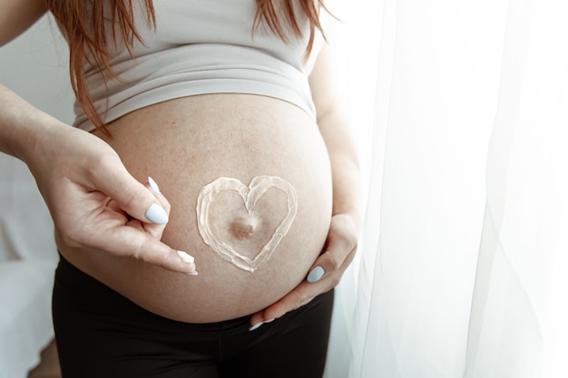Nahaufnahme des nackten Bauches einer zukünftigen Mutter in den letzten Monaten der Schwangerschaft mit einer bemalten Herzcreme.