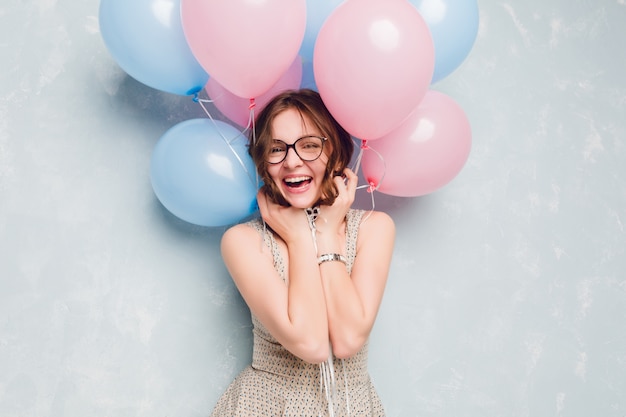 Nahaufnahme des niedlichen brünetten Mädchens, das in einem Studio steht, breit lächelt und mit blauen und rosa Luftballons spielt. Sie hat Spaß