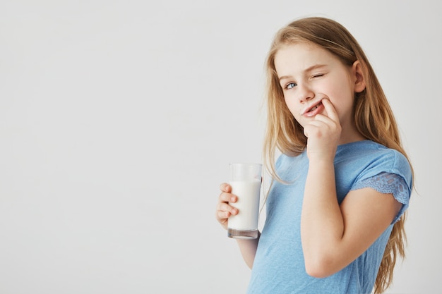 Nahaufnahme des niedlichen kleinen Mädchens mit den blauen Augen schaut zur Seite, trinkt Glas Milch und putzt die Zähne nach einer Mahlzeit mit dem Finger. Sorglose Kindheit.