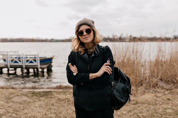 Nahaufnahme des Outdoor-Porträts eines selbstbewussten, stilvollen Mädchens mit Sonnenbrille, das eine Mütze und eine dunkle Jacke trägt und ihre Hände auf die Brust gegen den See mit Booten legt