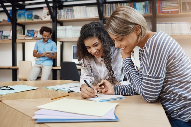 Nahaufnahme des Paares schöne junge multiethnische Studentinnen, die zusammen Hausaufgaben machen, Aufsatz für Präsentation schreiben, sich auf Prüfungen mit guter Laune vorbereiten