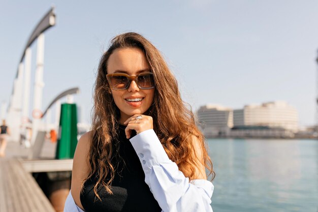 Nahaufnahme des Porträts des langhaarigen europäischen Modells mit Sonnenbrille und blauem Hemd, das vor der Kamera auf dem Hintergrund des Sees posiert Junge attraktive kaukasische Frau, die im Freien mit vollem Mund schwingende Haare lacht